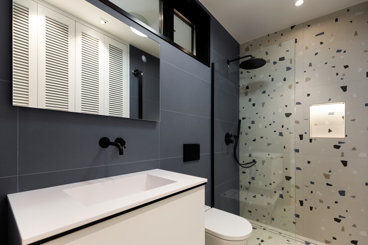 Baño con pared negra, lavabo blanco y ducha de terrazo en una vivienda reformada