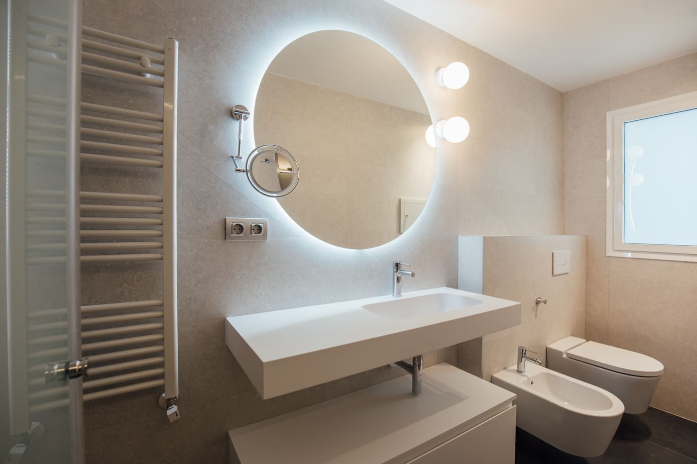Baño con espejo redondo iluminado y toallero tras la reforma