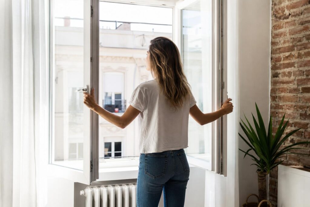 Woman opening large white windows in a bright room with an exposed brick wall to ventilate her apartment.