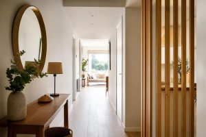 Bright minimalist hallway with a wooden console table, round mirror, and vertical wood slats leading to a sunlit living room.