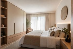 Modern minimalist bedroom in beige and wood tones featuring a large built-in wardrobe, a plush bed with neutral linens, and soft natural light.