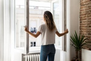 Woman opening large white windows in a bright room with an exposed brick wall to ventilate her apartment.